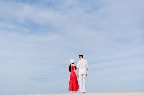 Free Asian couple in red dress and white outfit holding hands under a bright blue sky. Stock Photo