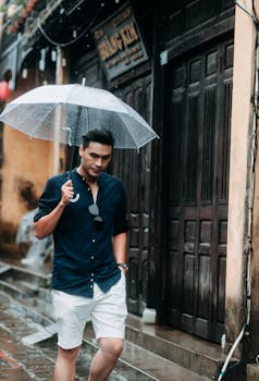 A stylish young man walks on a rainy street holding a transparent umbrella, wearing casual attire.