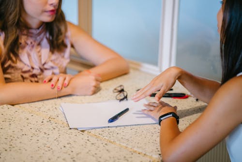 Two persons sitting across each other. There is a table in between them with a pair of glasses, paper, and pens.