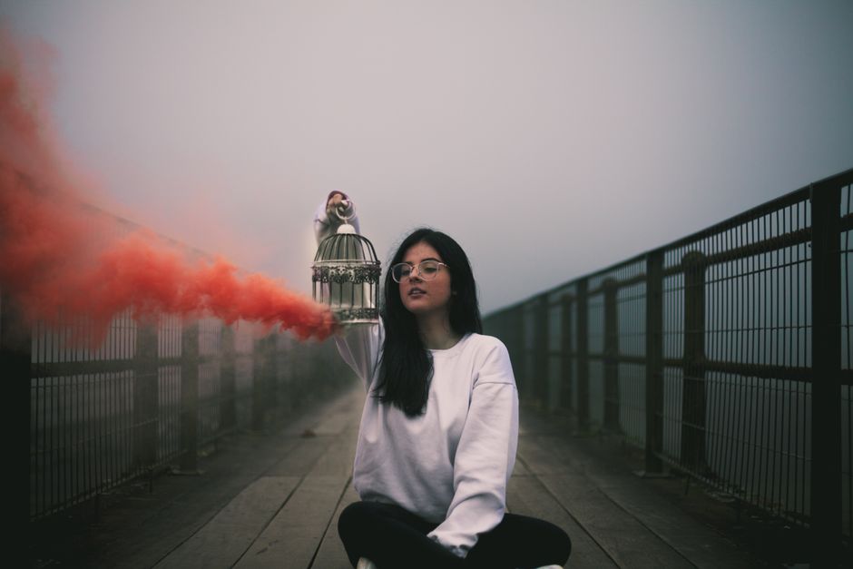A woman sits on a foggy bridge holding a birdcage with orange smoke, in Paranapiacaba.