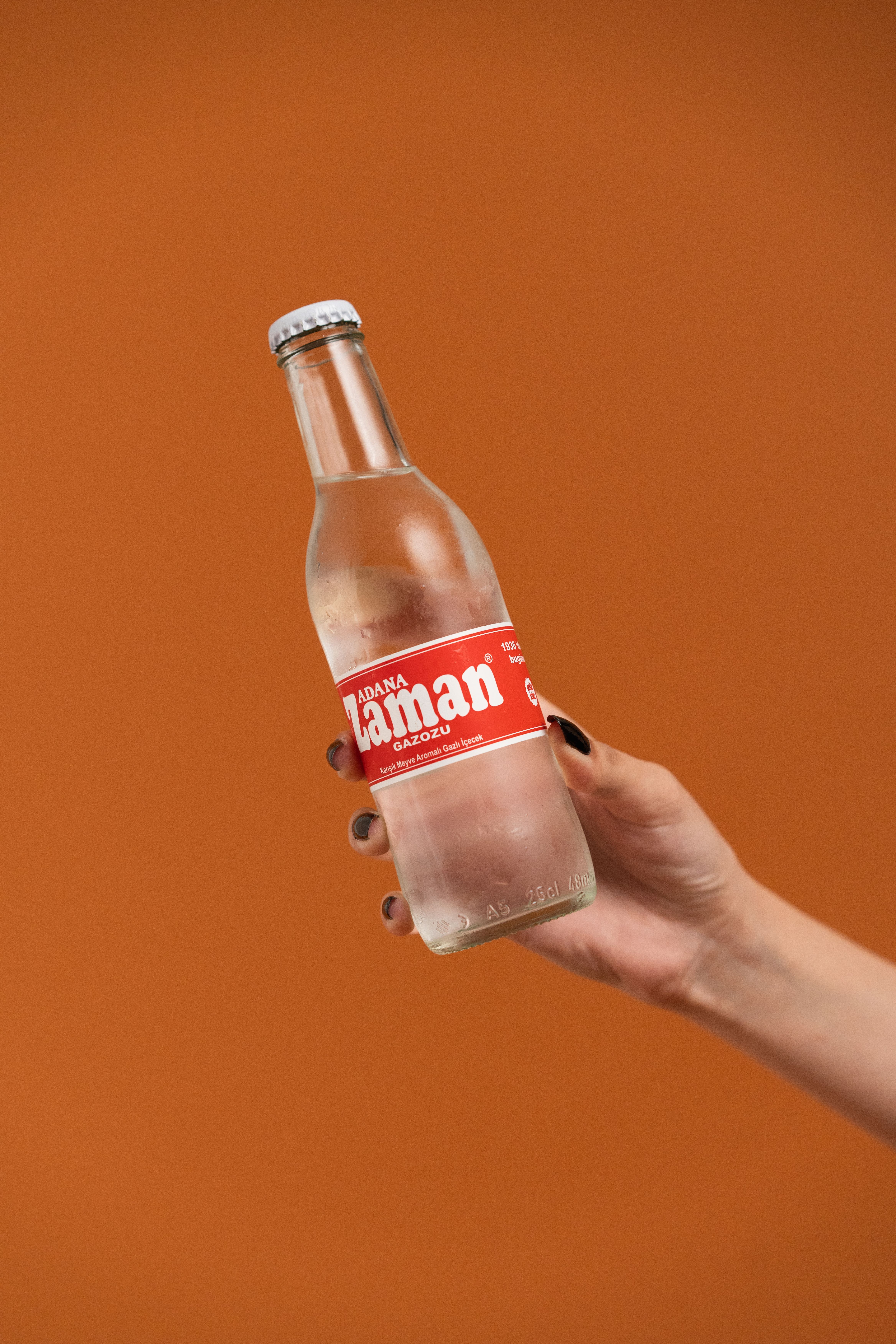 A hand holds a chilled soda bottle with a red label, set against an orange background.