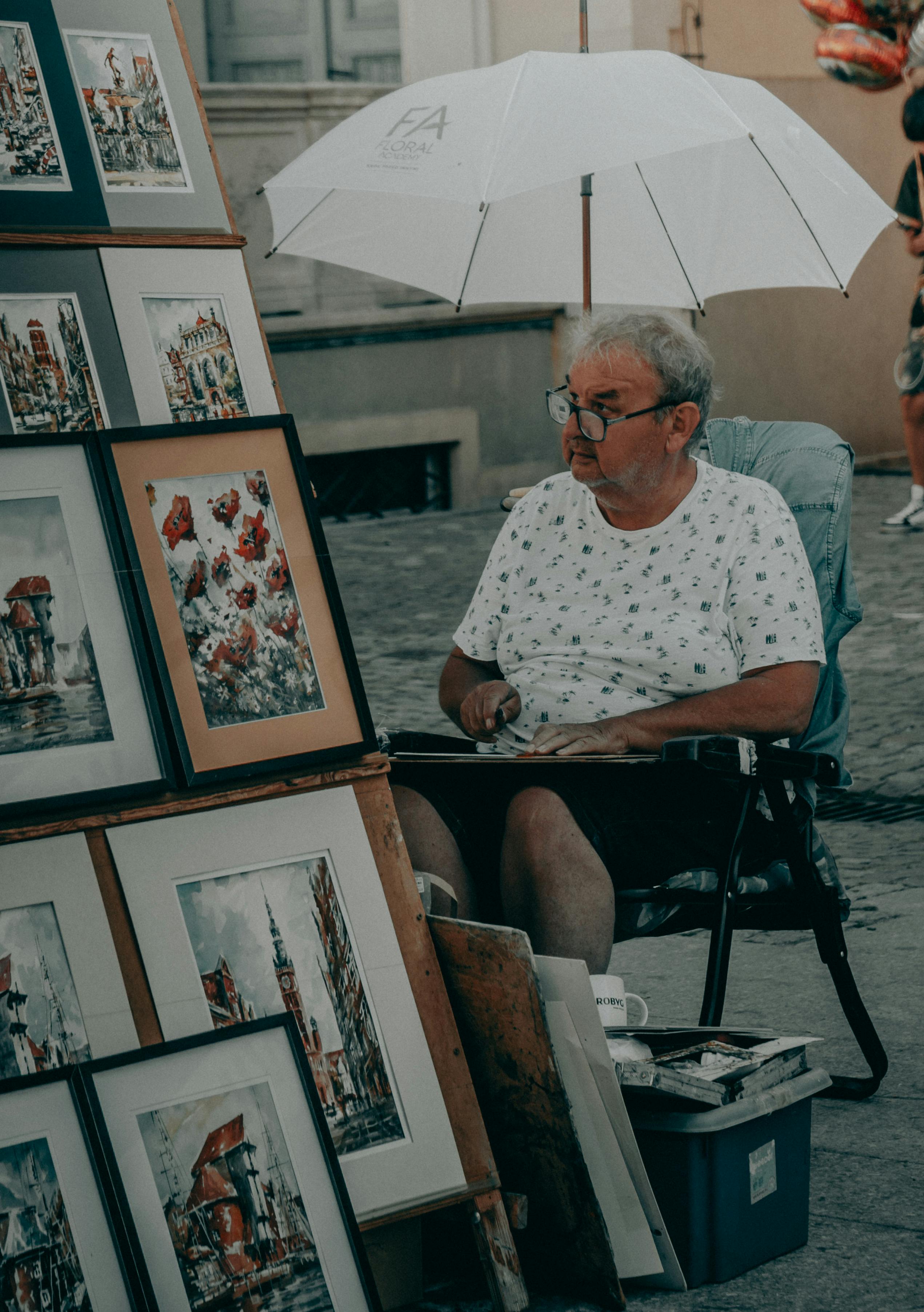 Senior man selling vibrant paintings at a street market, sitting under a white umbrella.