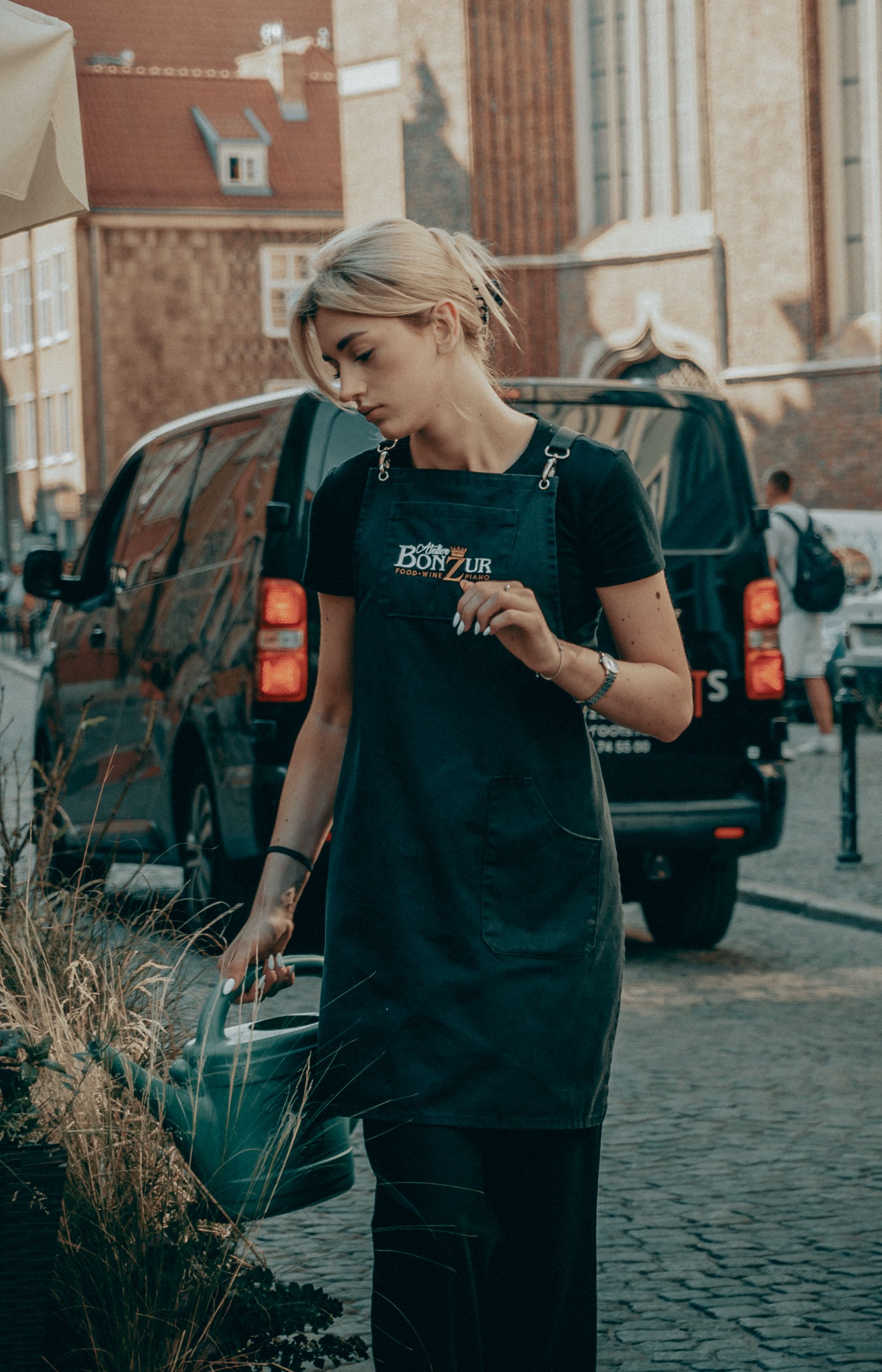 A young blonde woman in urban attire waters plants with a watering can on a city street.