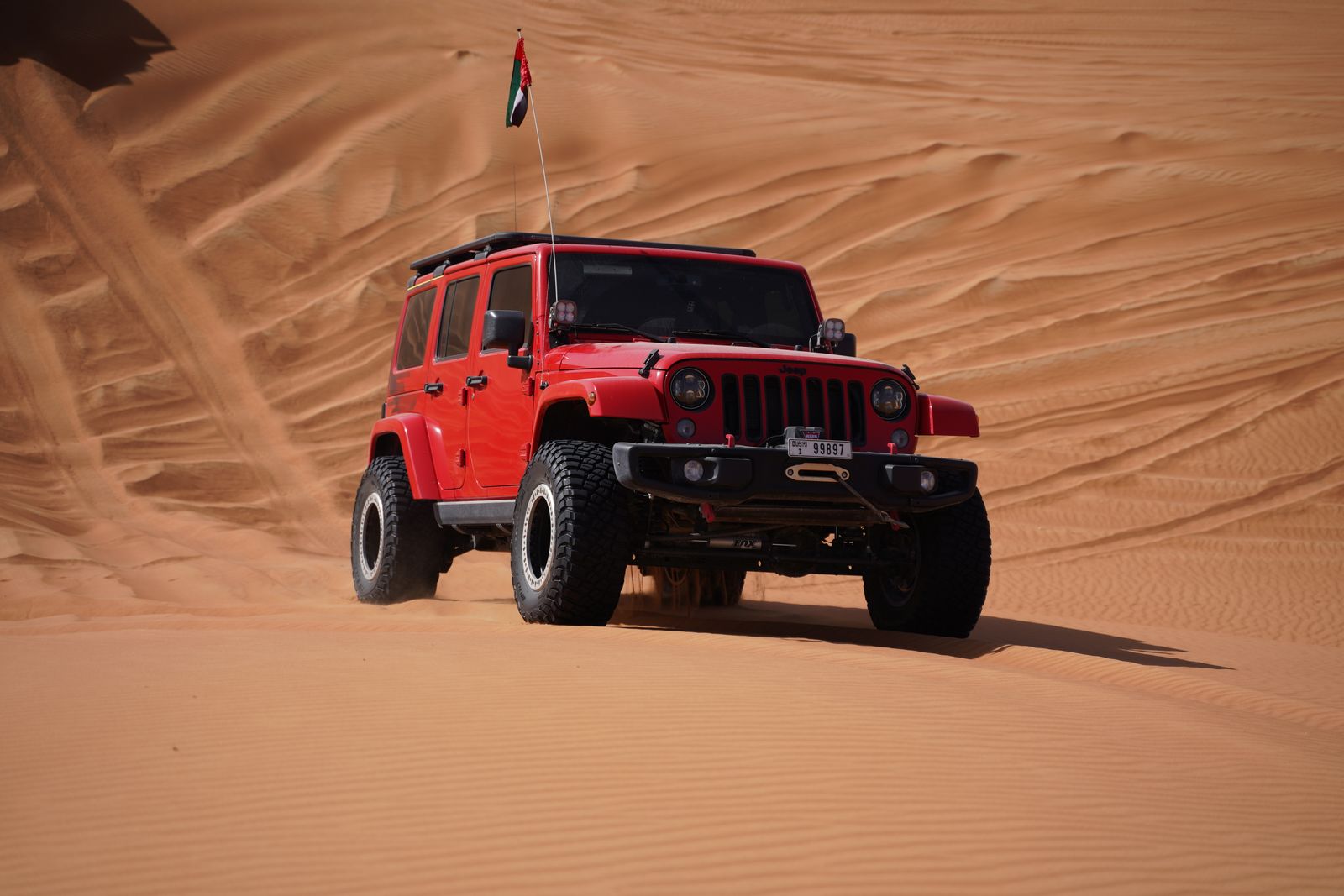 Red Jeep with aired-down off-road tires parked on red sand dunes at sunset
