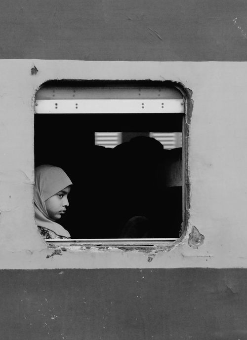 Free A young girl wearing a hijab in a black and white portrait, looking out from a train window. Stock Photo