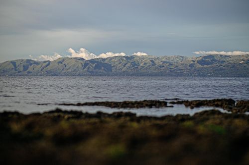 Free Tranquil coastal scene with mountains reflecting in the ocean under a cloudy blue sky. Stock Photo