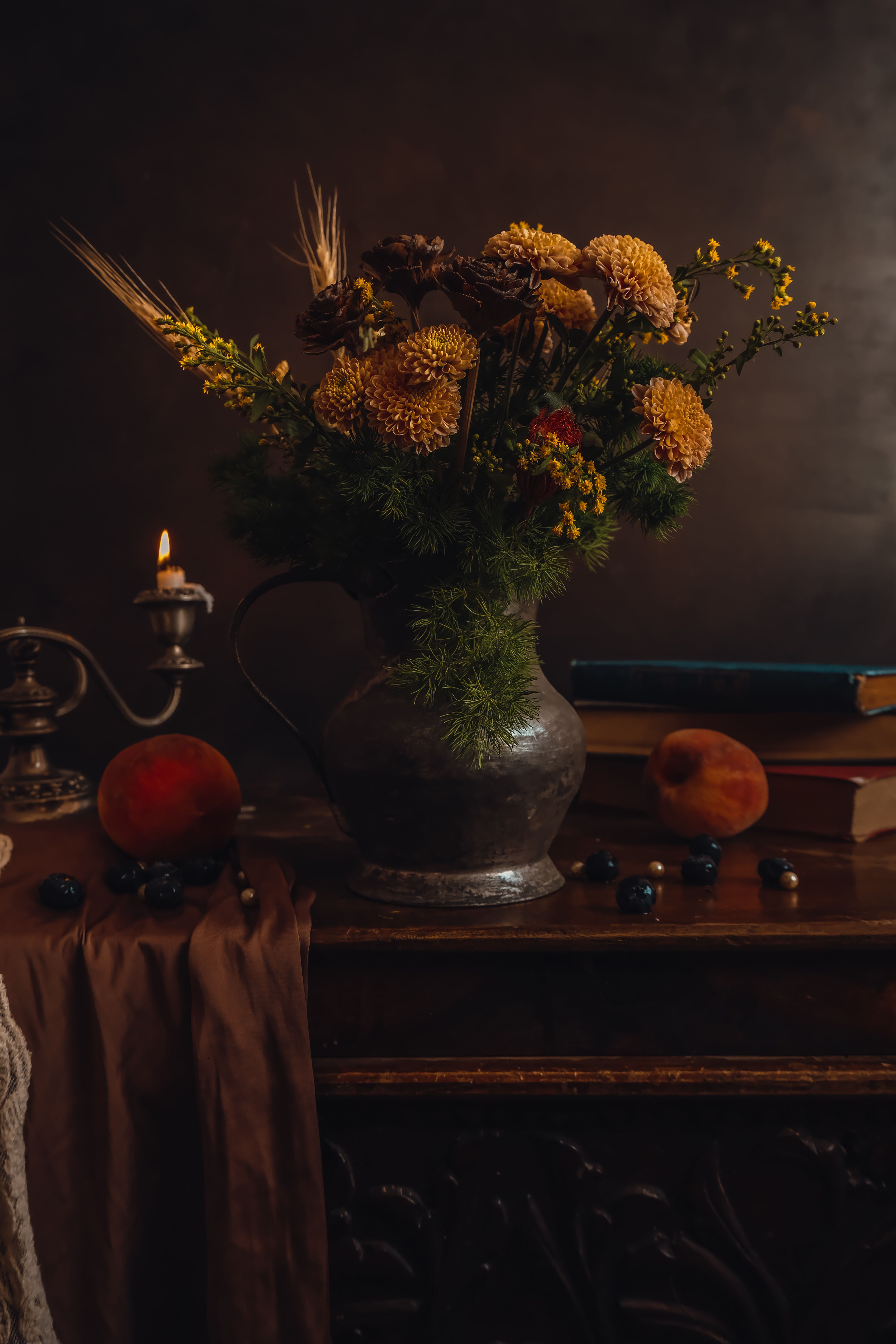 Elegant still life with yellow flowers, antique metal jug, and books on wooden cabinet with candlelight.