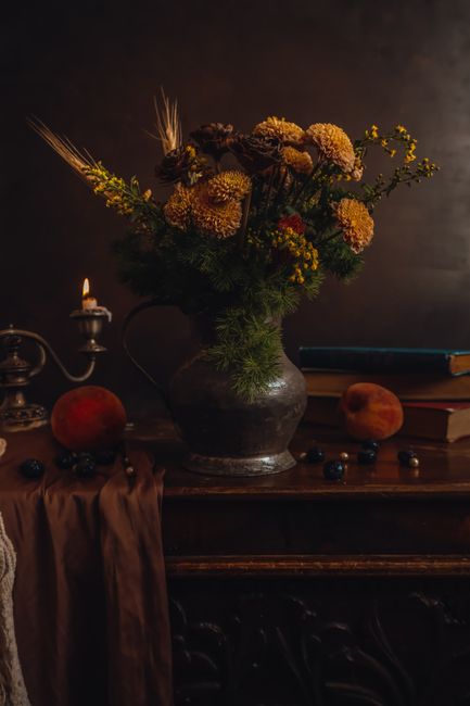 Elegant still life with yellow flowers, antique metal jug, and books on wooden cabinet with candlelight.