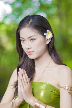 Asian woman with a flower in her hair, hands clasped, wearing a traditional green dress in a serene outdoor setting.