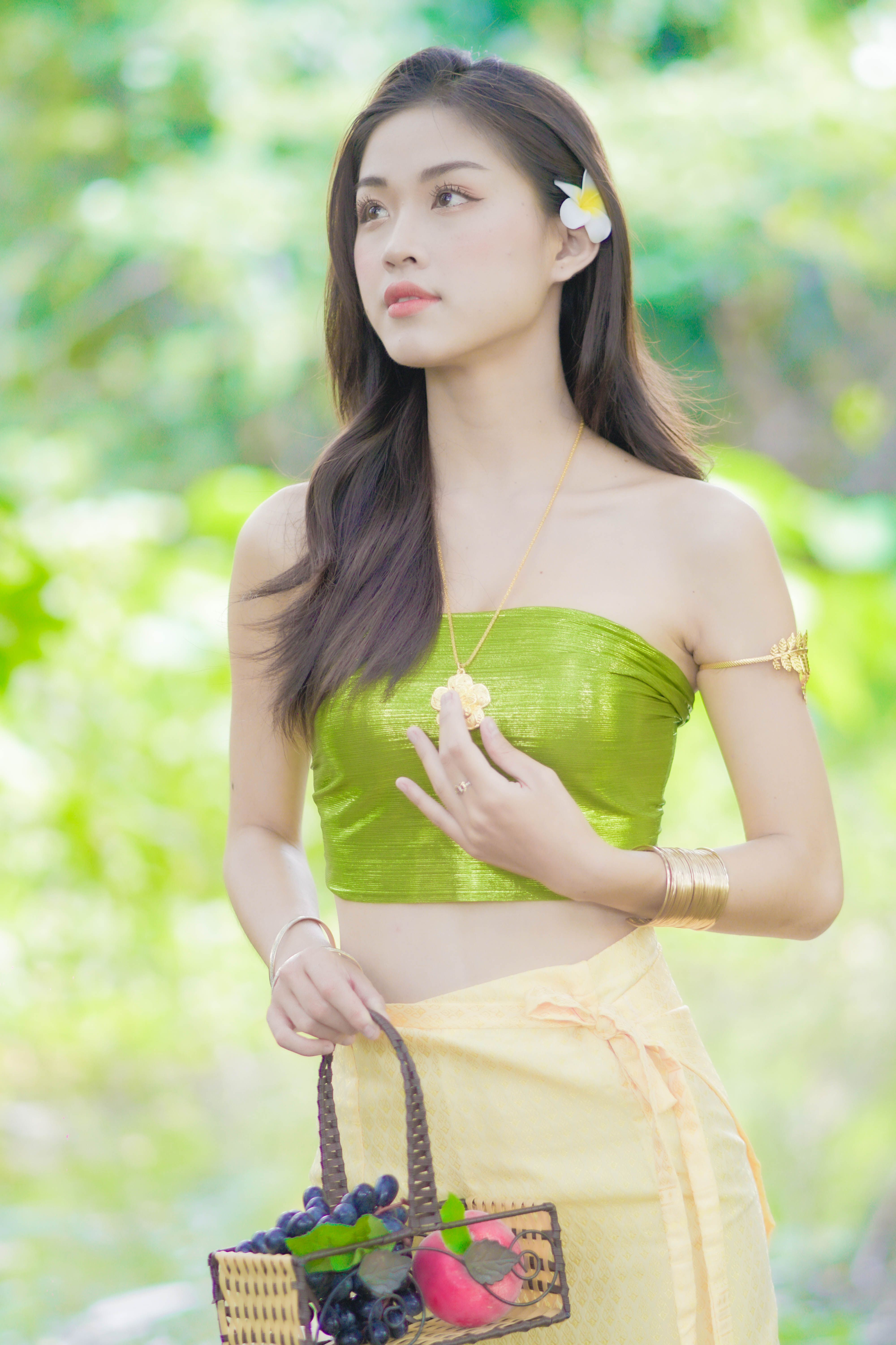 A beautiful woman in traditional attire holding a basket of fruits in a lush outdoor setting.