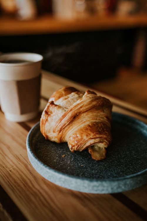 Free Warm croissant on a ceramic plate with steaming coffee in a cozy café setting. Stock Photo