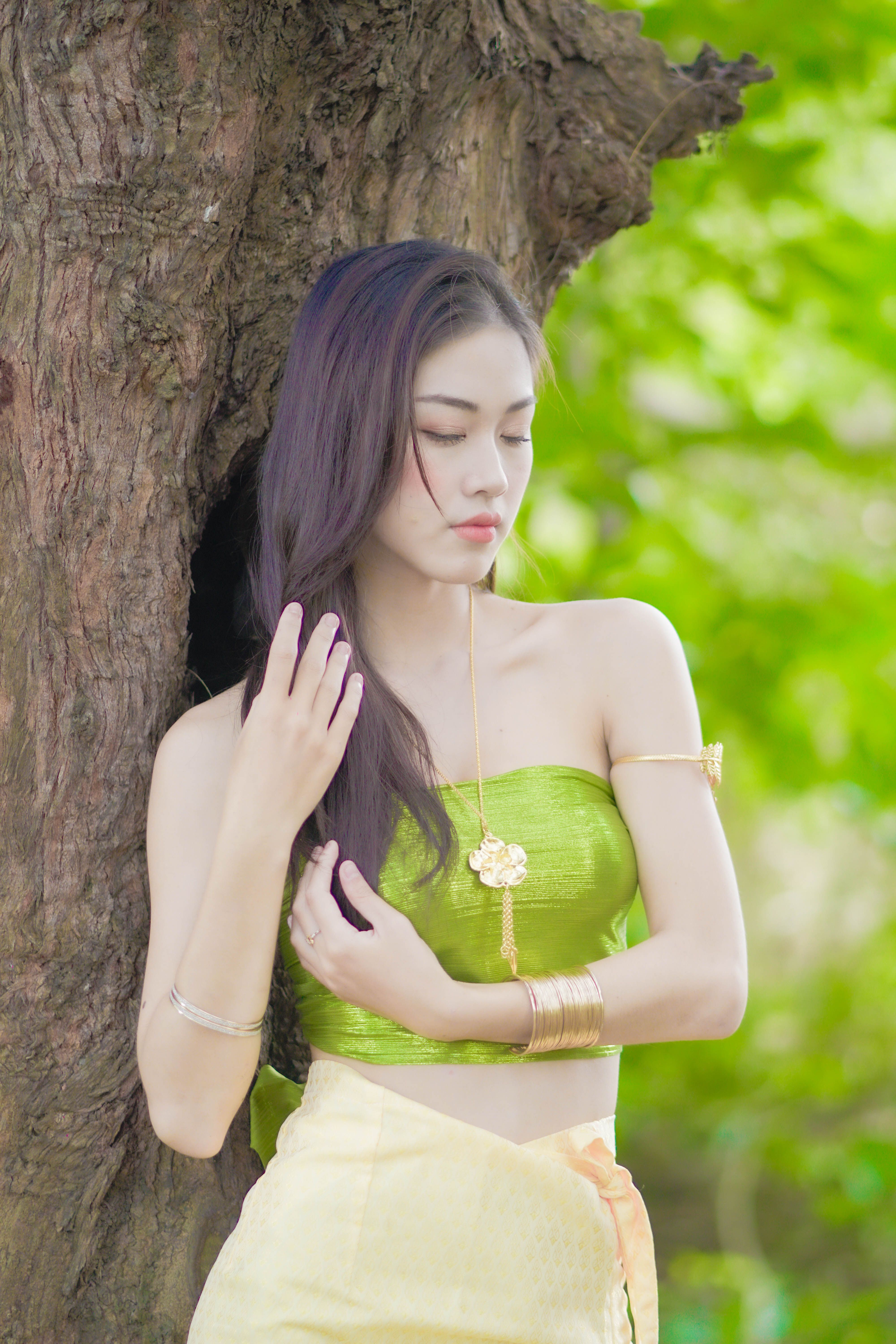 Asian woman in green tube top with jewelry poses by tree trunk in serene outdoor setting.