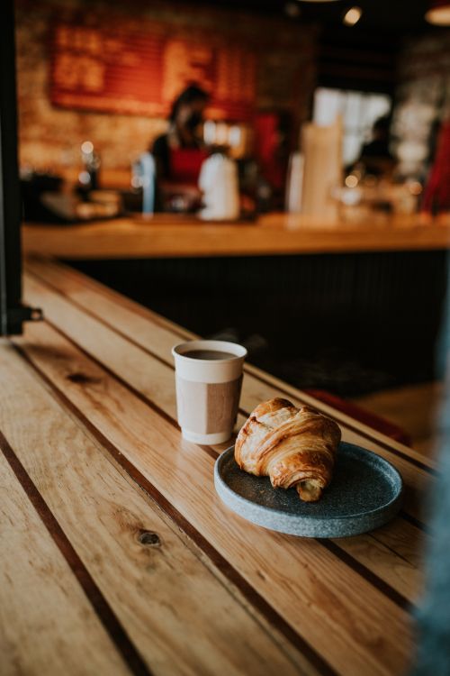 Free Warm café scene featuring a croissant and cup of coffee on a wooden table, perfect for breakfast imagery. Stock Photo