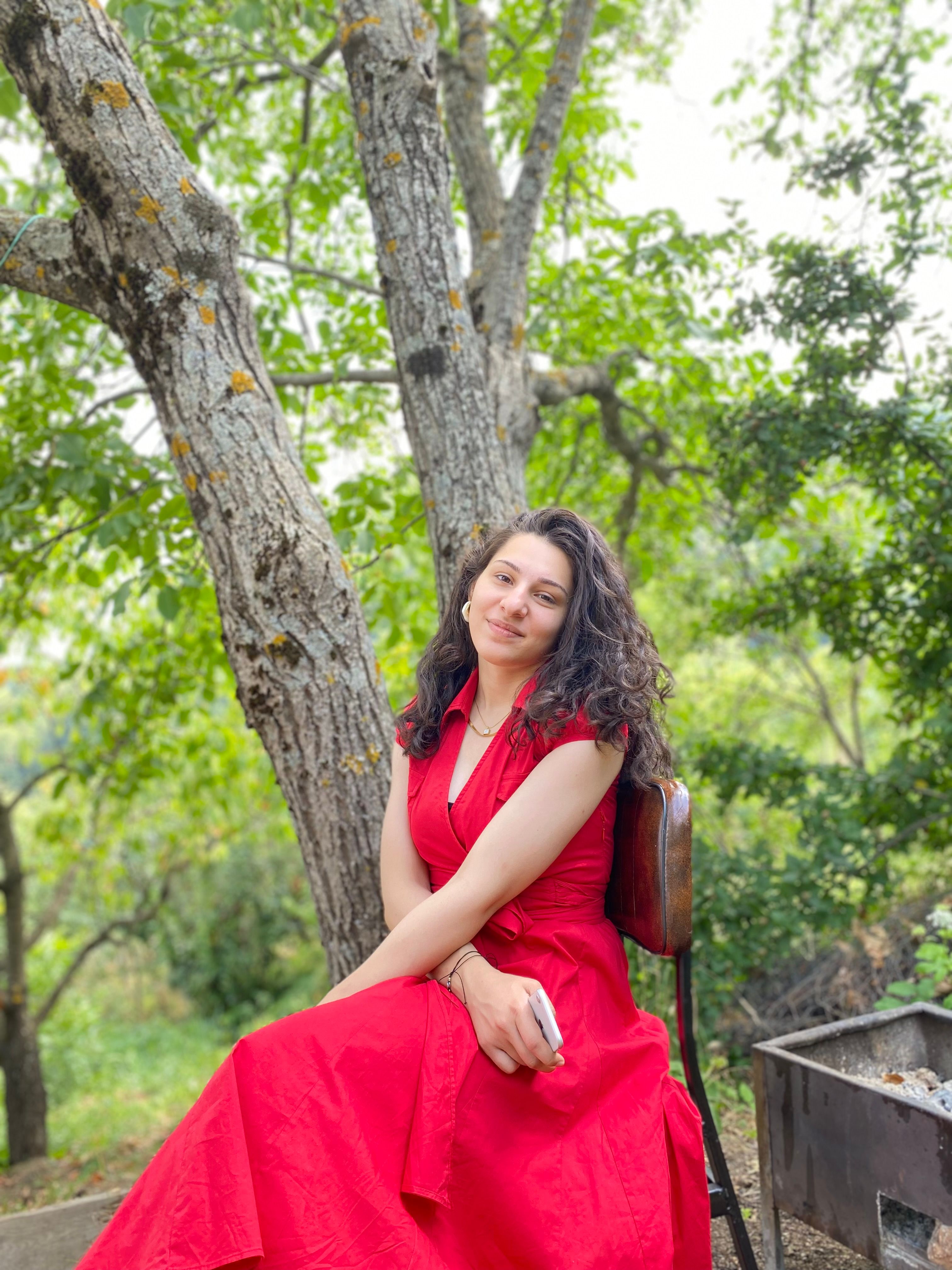 A smiling woman in a sleeveless red dress sits on a chair under a tree in Bursa, Turkey.