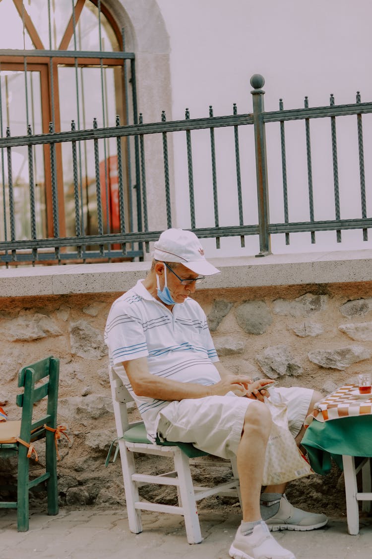 Man In White Polo Shirt Wearing Hat Using Phone  While Sitting On Chair 