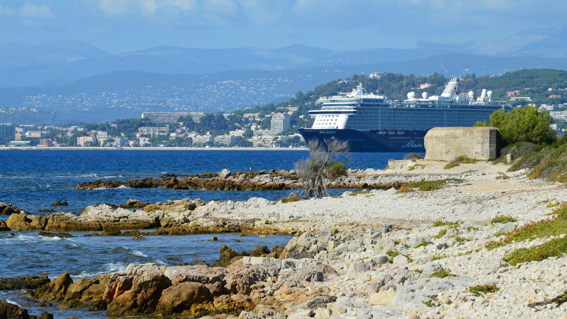 A luxury cruise ship sails along a scenic rocky coastline with a city skyline backdrop.