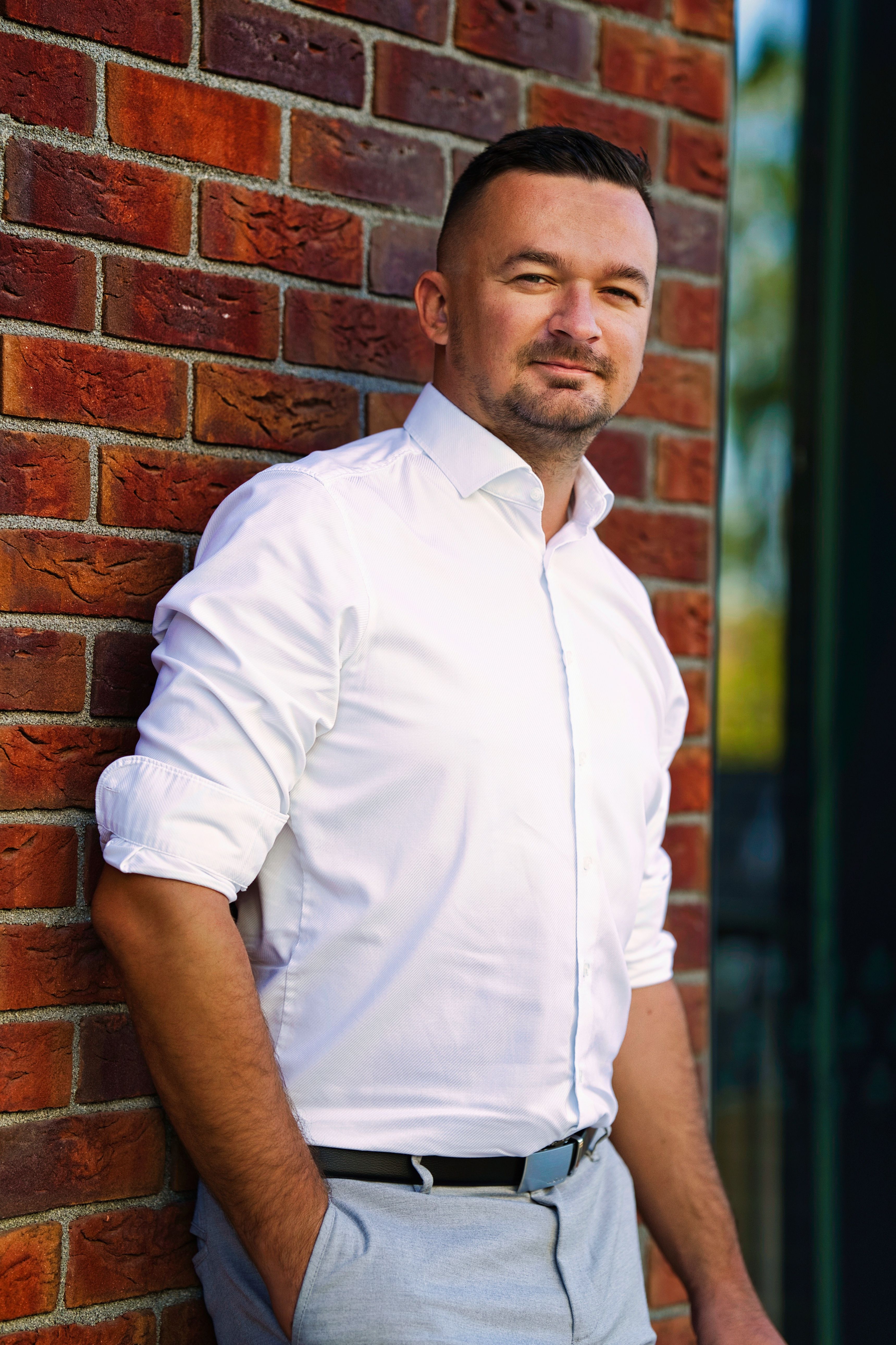 Caucasian man confidently posing against a sunlit brick wall.