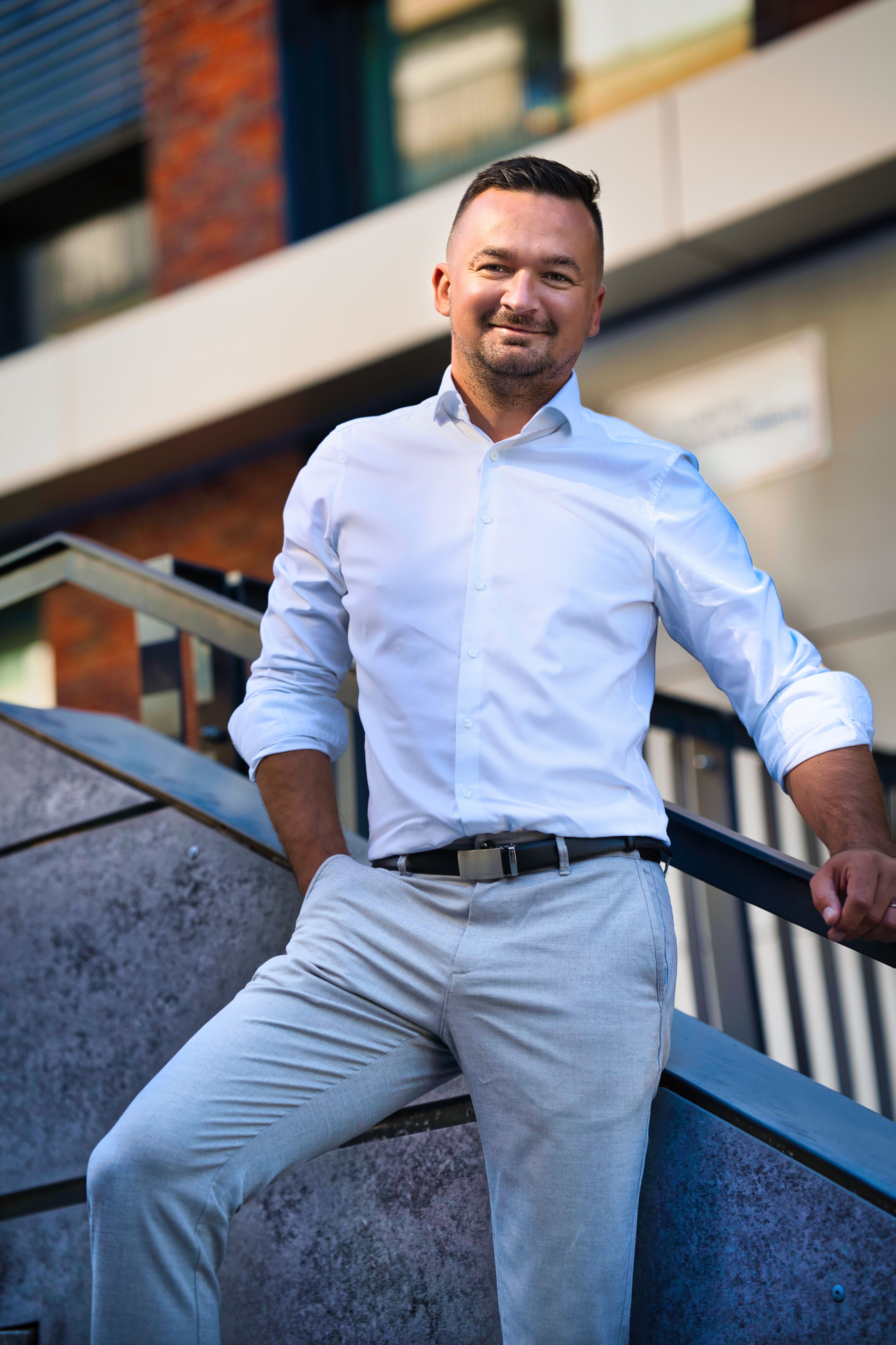 Confident man posing on a modern staircase in Bratislava, wearing a white shirt and light gray pants.