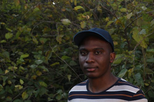 Man in Striped Shirt and Cap Standing Near Green Leaves