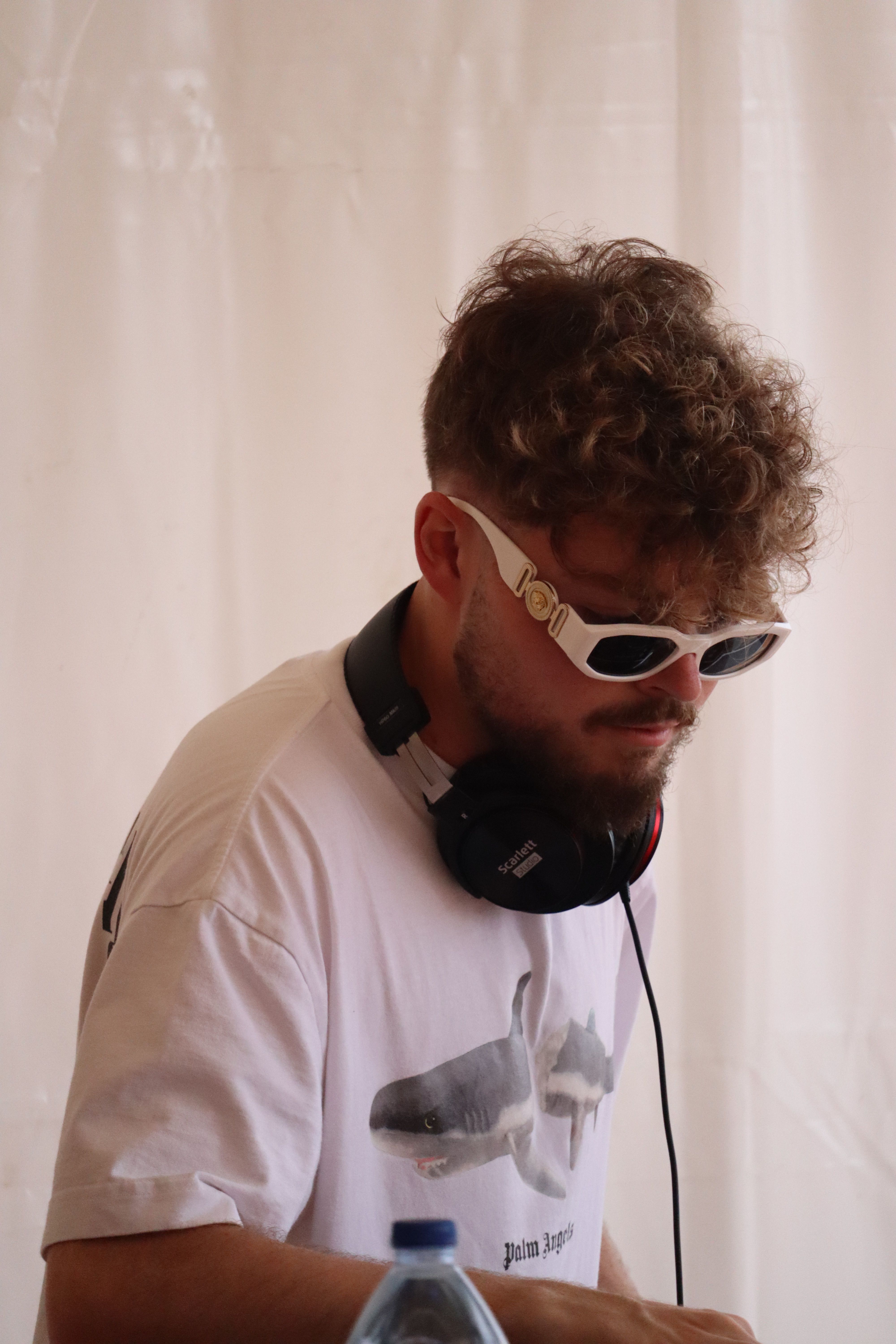 DJ with curly hair and sunglasses wearing headphones, focused on mixing music.