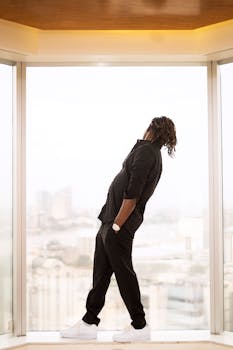 Man in black attire standing at a large window looking at urban scenery outside.