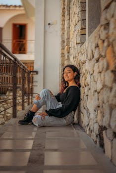 A young woman in ripped jeans and wavy hair relaxes on a stone balcony, exuding style and warmth.