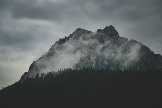Dramatic mountain landscape with fog and forest silhouettes under cloudy skies.