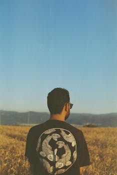 A man with a design shirt stands in a field under a clear blue sky, facing away.