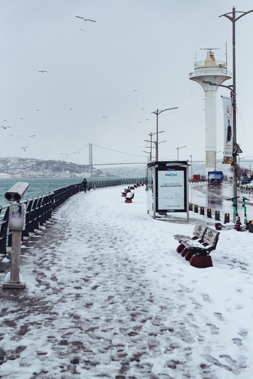 Free Serene snowy seaside promenade with benches facing a distant bridge on a winter day. Stock Photo