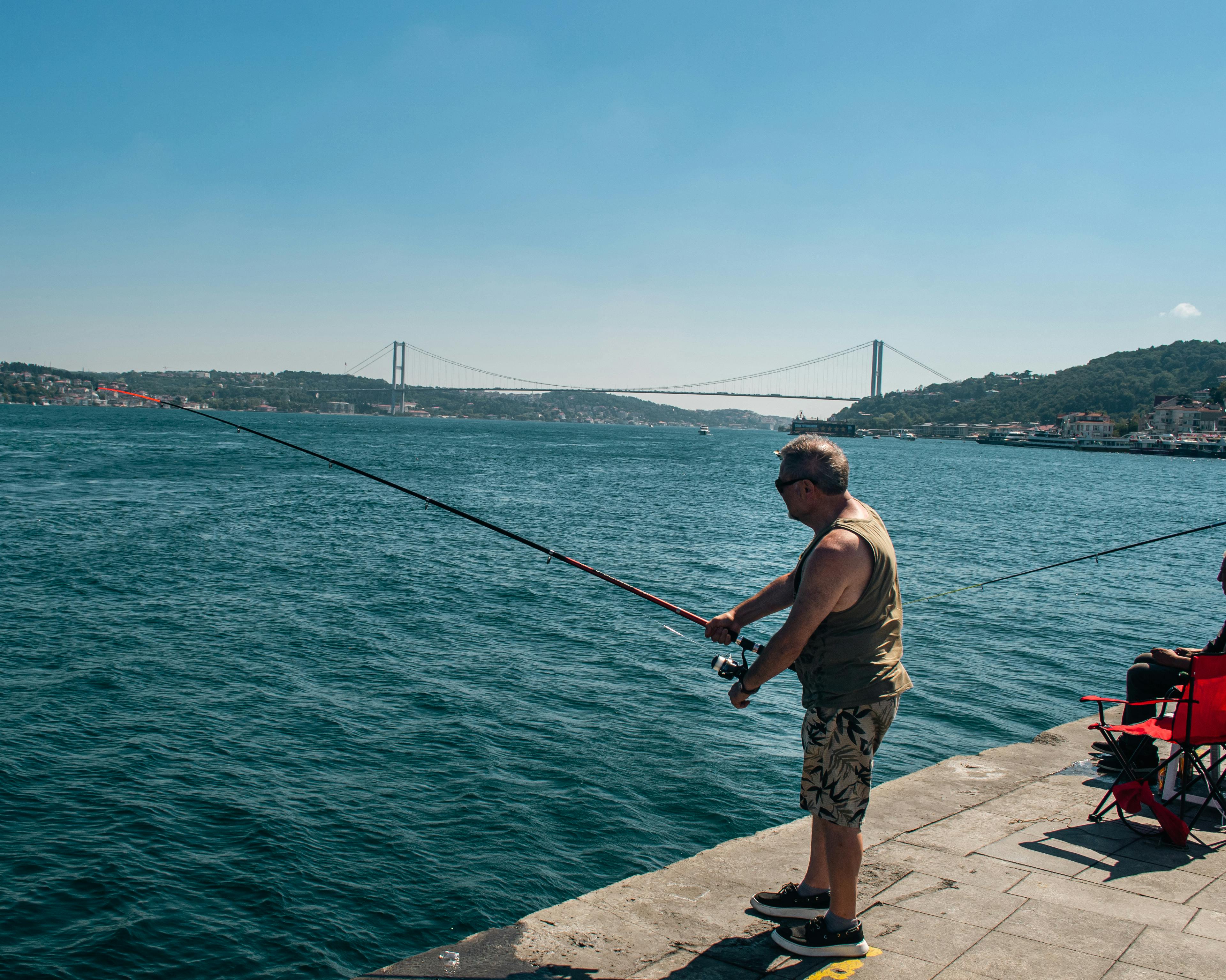 A man fishes at the edge of the Bosphorus in Istanbul with a scenic view of the bridge.