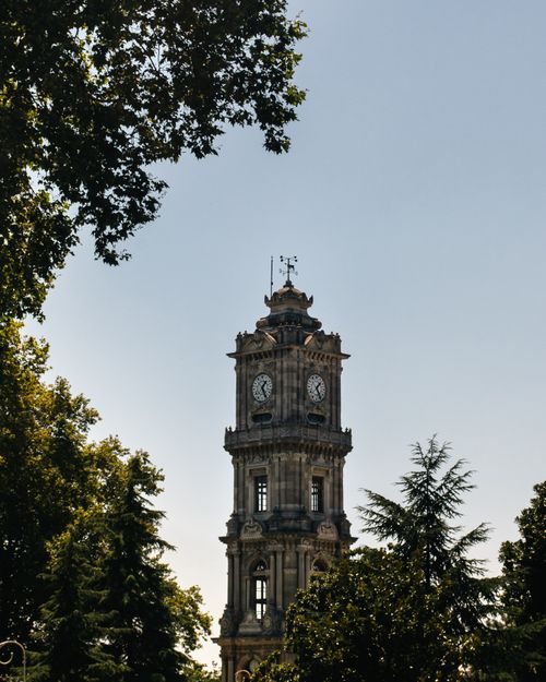 Free A beautiful view of the Dolmabahçe clock tower in Istanbul surrounded by lush green trees under a clear blue sky. Stock Photo