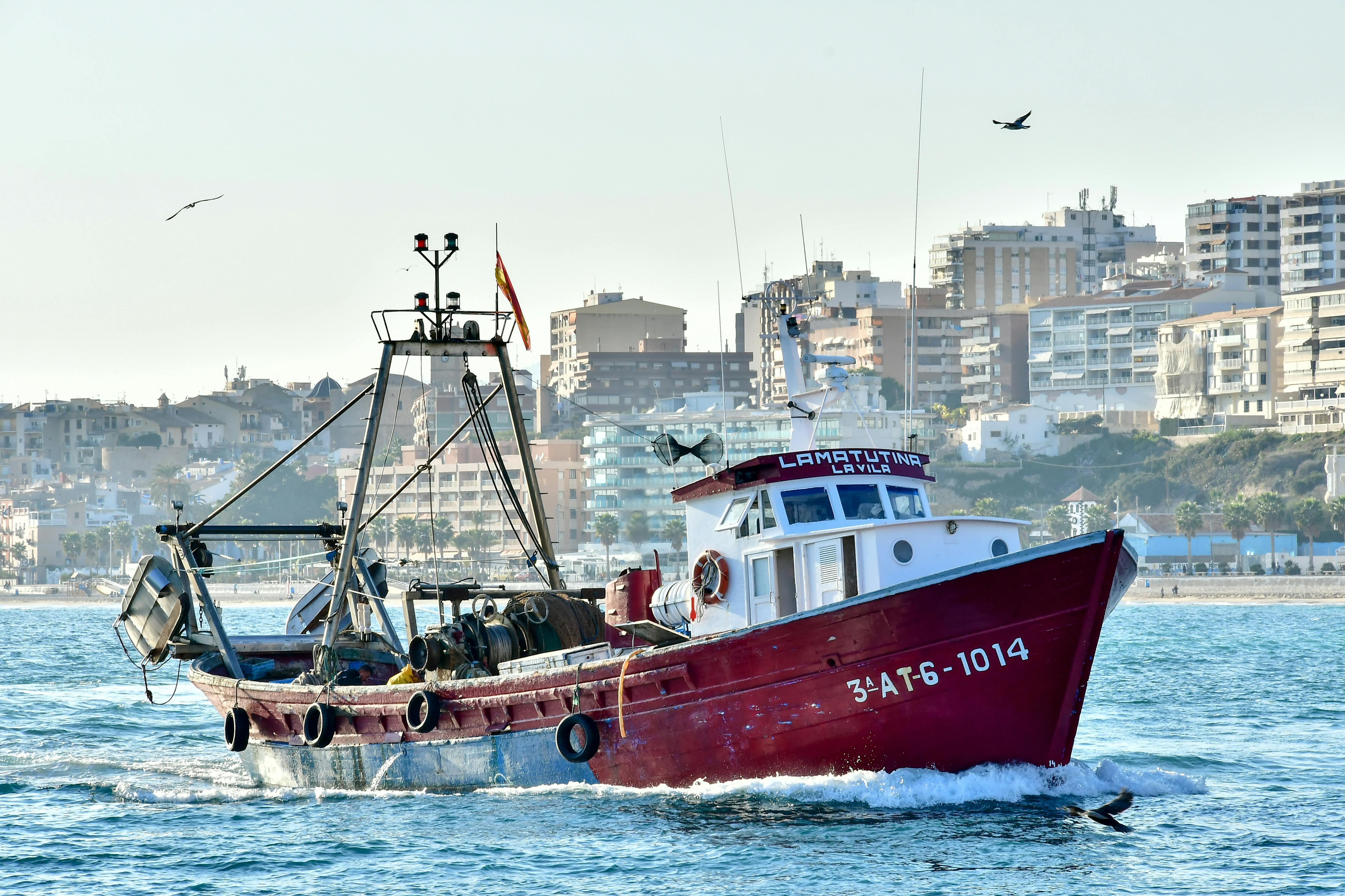 Free Red fishing boat cruising through Villajoyosa, Spain's waters with cityscape backdrop. Stock Photo