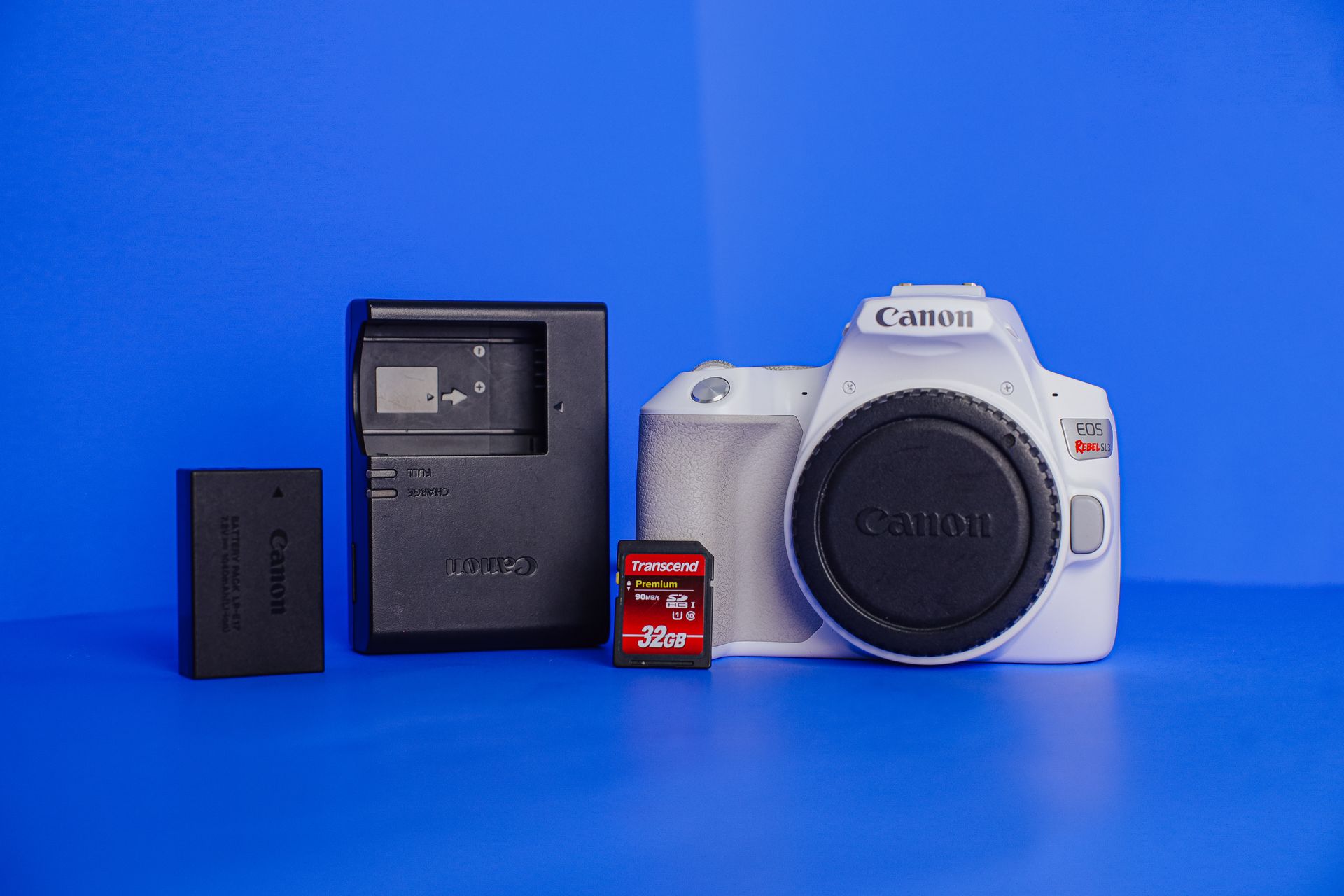 White camera, battery charger, and 32GB memory card on vibrant blue background.