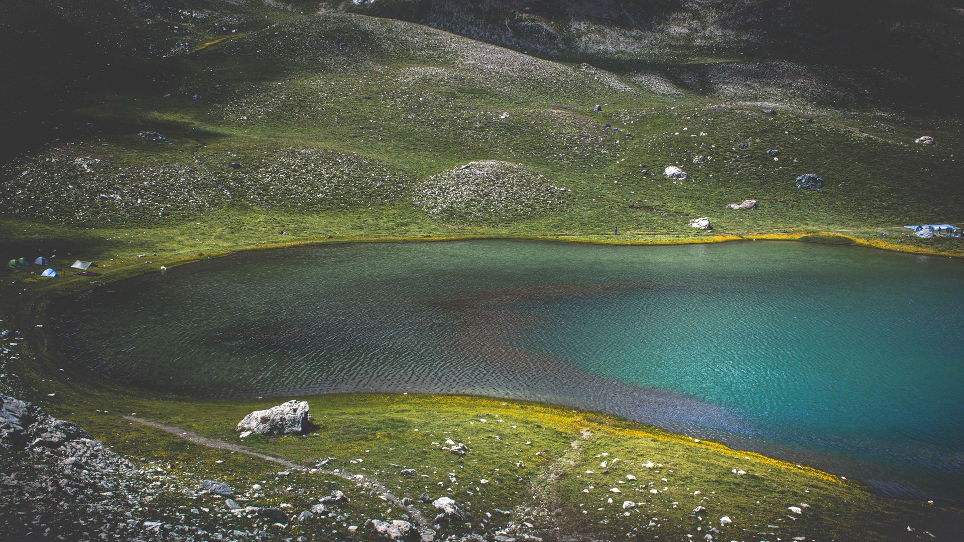A tranquil emerald lake surrounded by lush green hills and rocks under the open sky.