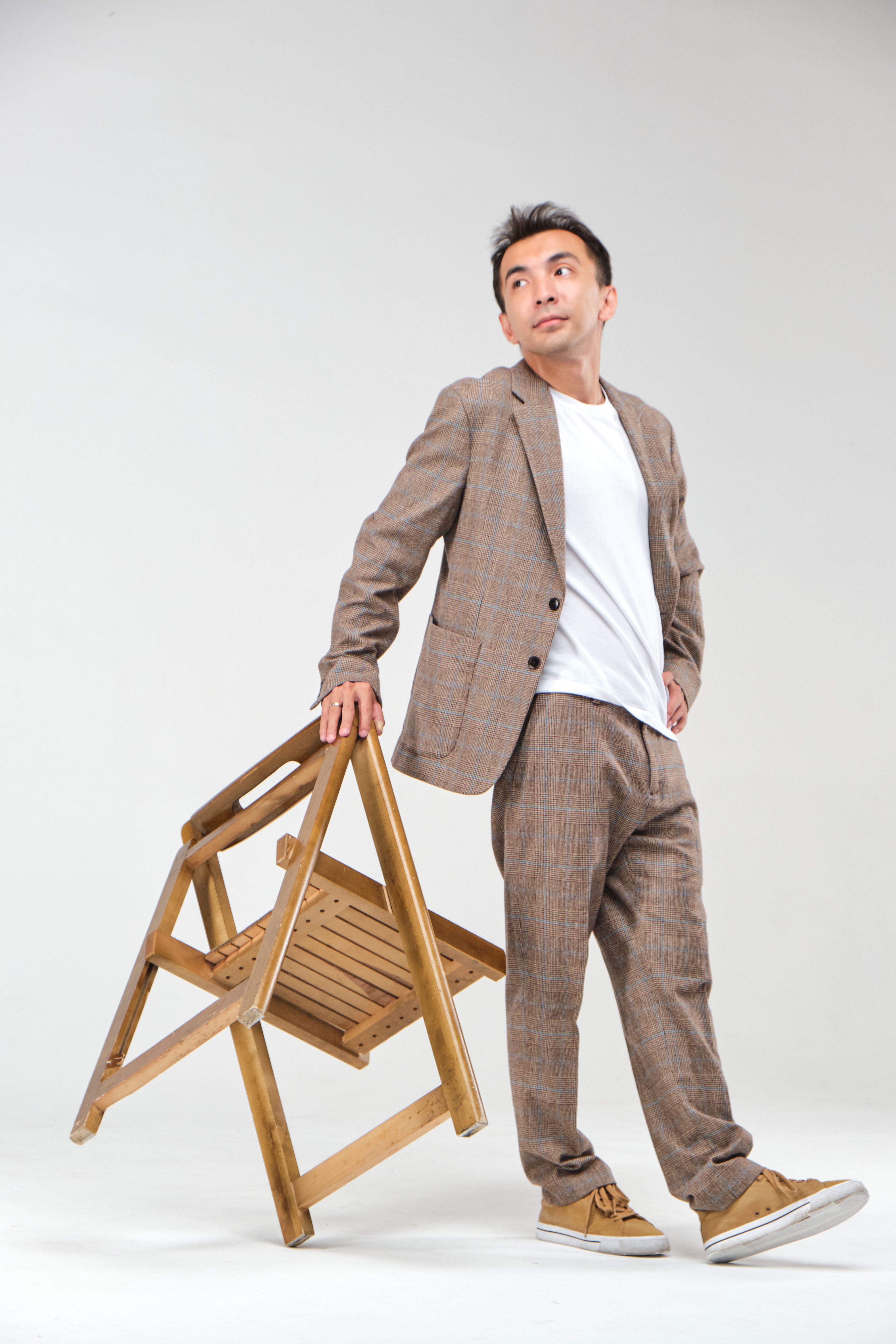 Man in casual suit posing with a folding chair in studio setting.