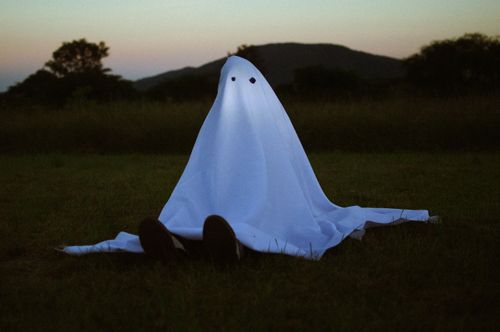Free A person in a ghost costume sits on a field during dusk, creating an eerie atmosphere. Stock Photo
