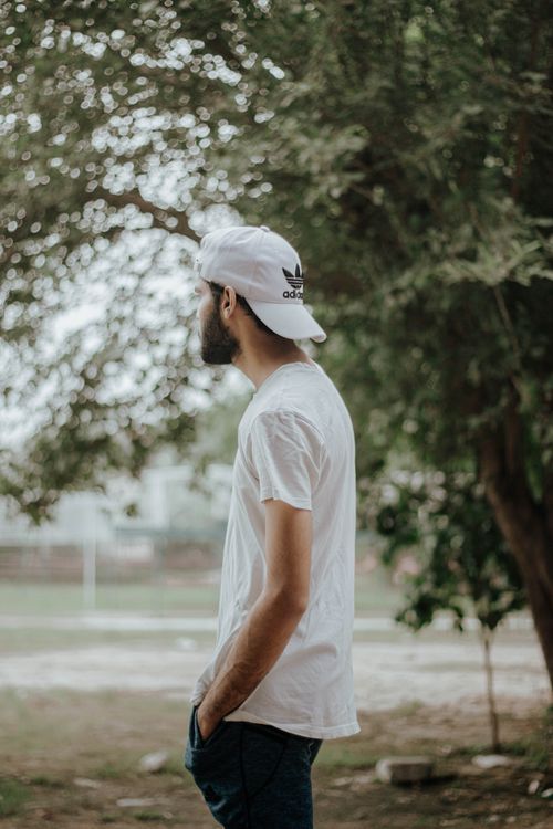 Free Side view of a man in a white shirt and cap standing outdoors with blurred background. Stock Photo
