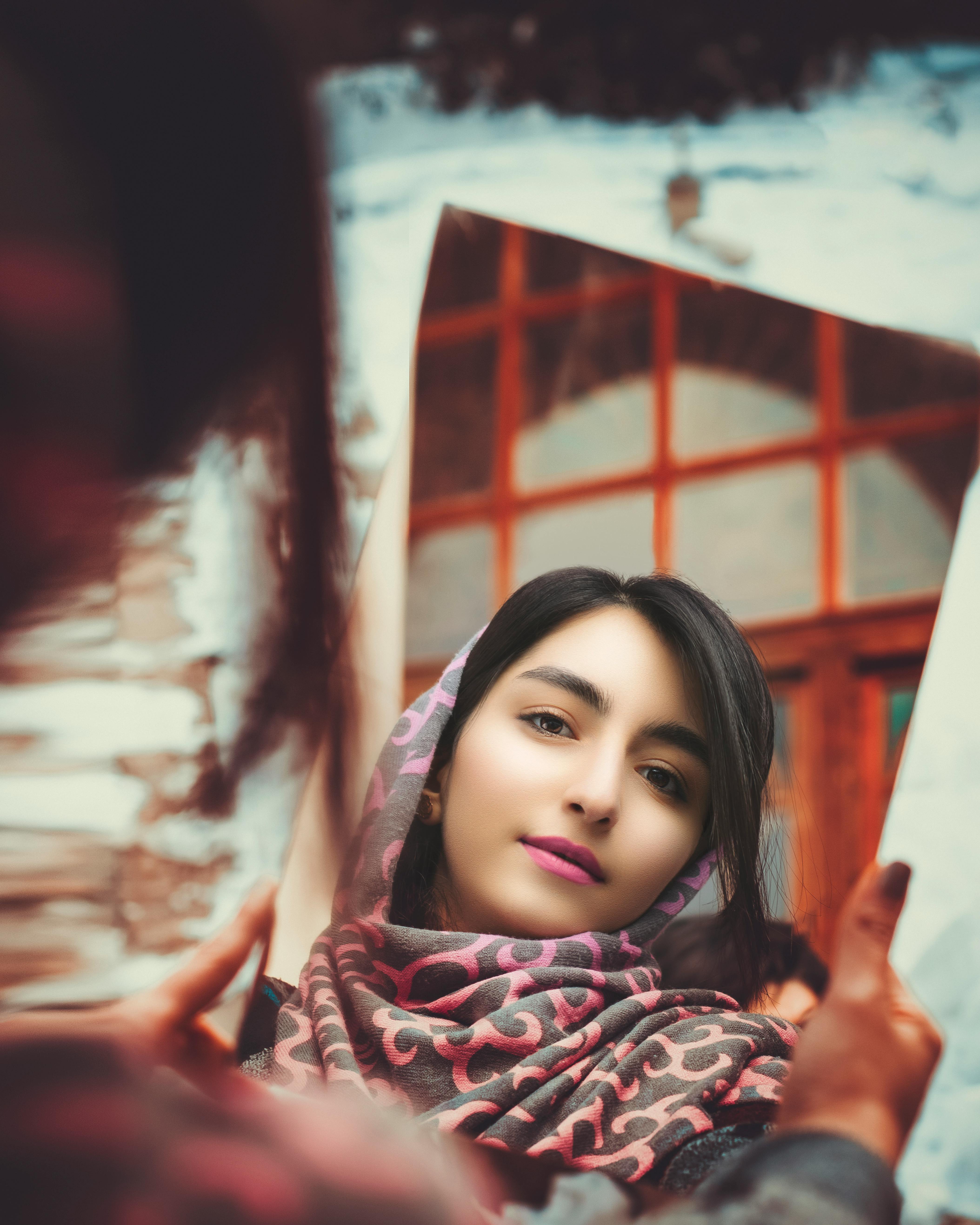 A young woman in a patterned scarf reflects in a mirror, capturing a vibrant outdoor scene.