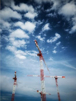 Three tower cranes stand tall against a vibrant blue sky with scattered clouds, showcasing construction progress.