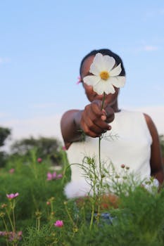 A person holding a white cosmos flower in a lush green outdoor setting.