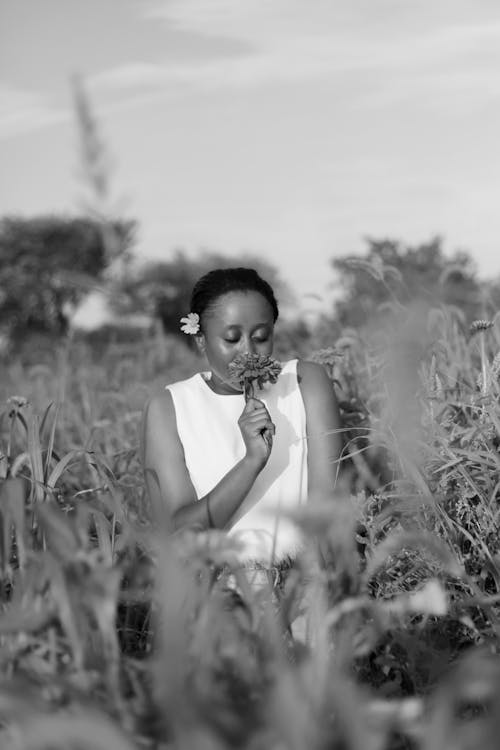Grayscale Photo of a Woman in a Field Smelling a Flower
