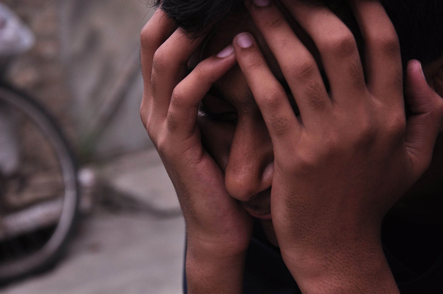 A close-up of a young man looking stressed, resting his face in his hands outdoors.