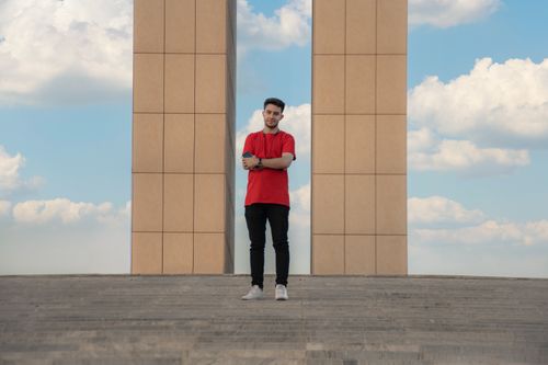 Free A man in a red shirt stands with arms crossed in front of a modern structure under a cloudy sky. Stock Photo