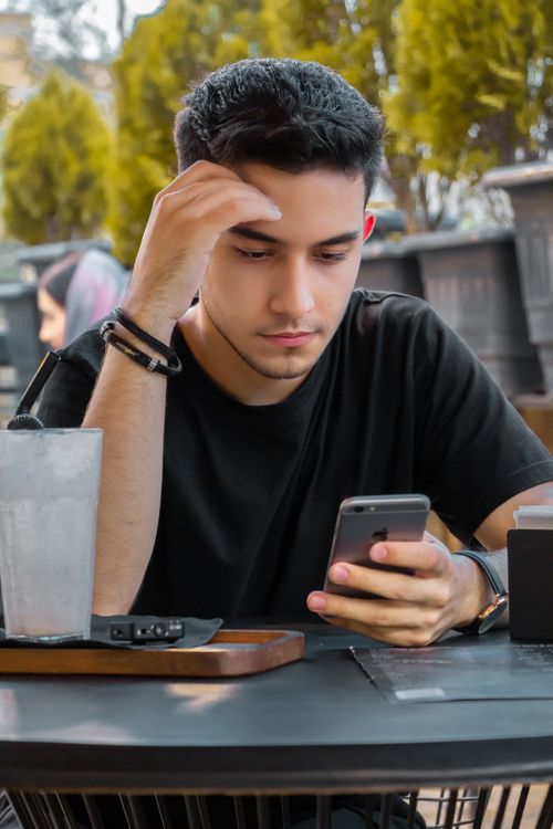 Free A young man in a black t-shirt, focused on his smartphone while seated at an outdoor table. Stock Photo