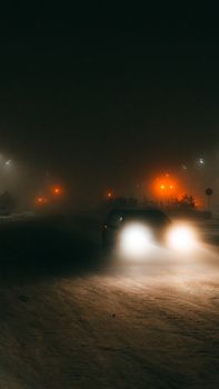 A car drives through snowy streets at night with glowing headlights and streetlights.