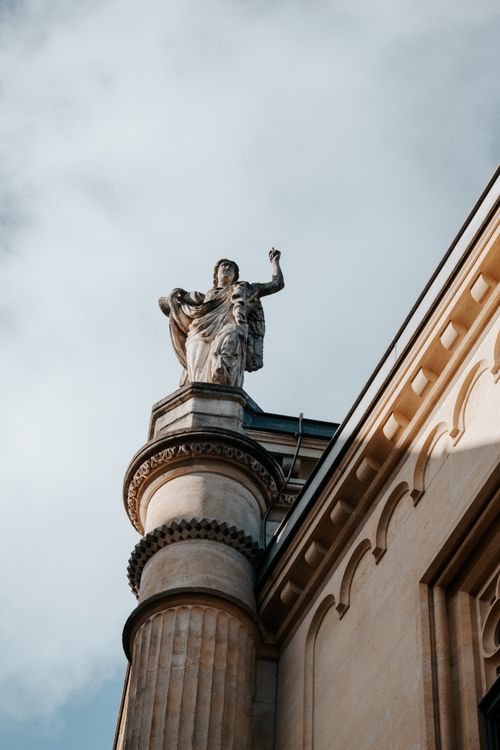 Free A classic stone statue stands atop an ornate marble column against a cloudy sky. Stock Photo