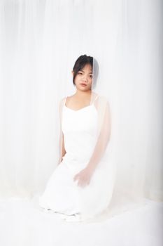 A young woman in a white dress sits gracefully behind sheer white curtains, creating a serene and elegant portrait.
