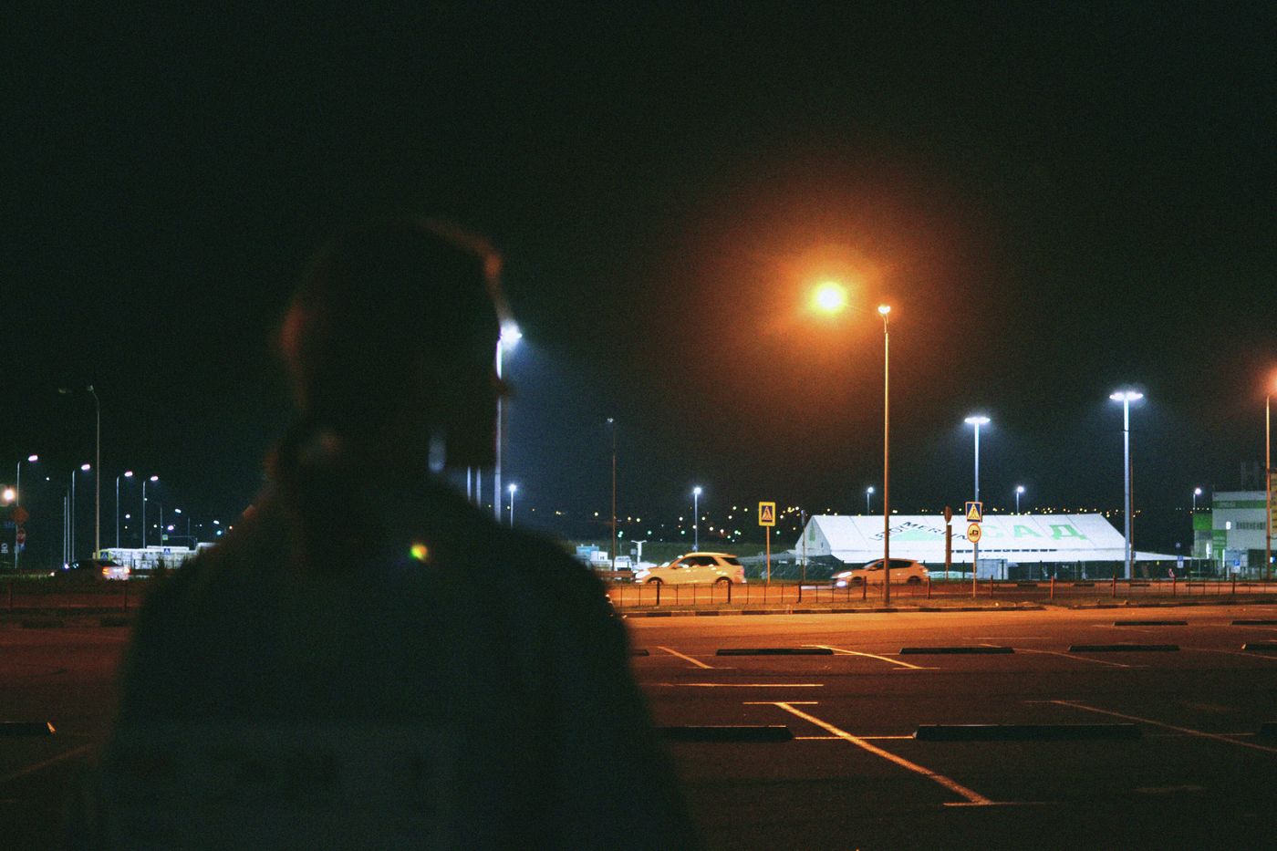 A person standing alone in a parking lot at night, city lights in the distance