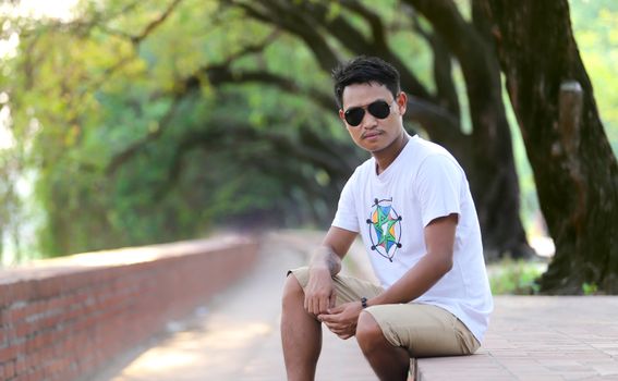 Asian man in sunglasses sitting casually on a pathway aligned with trees, enjoying a sunny day.
