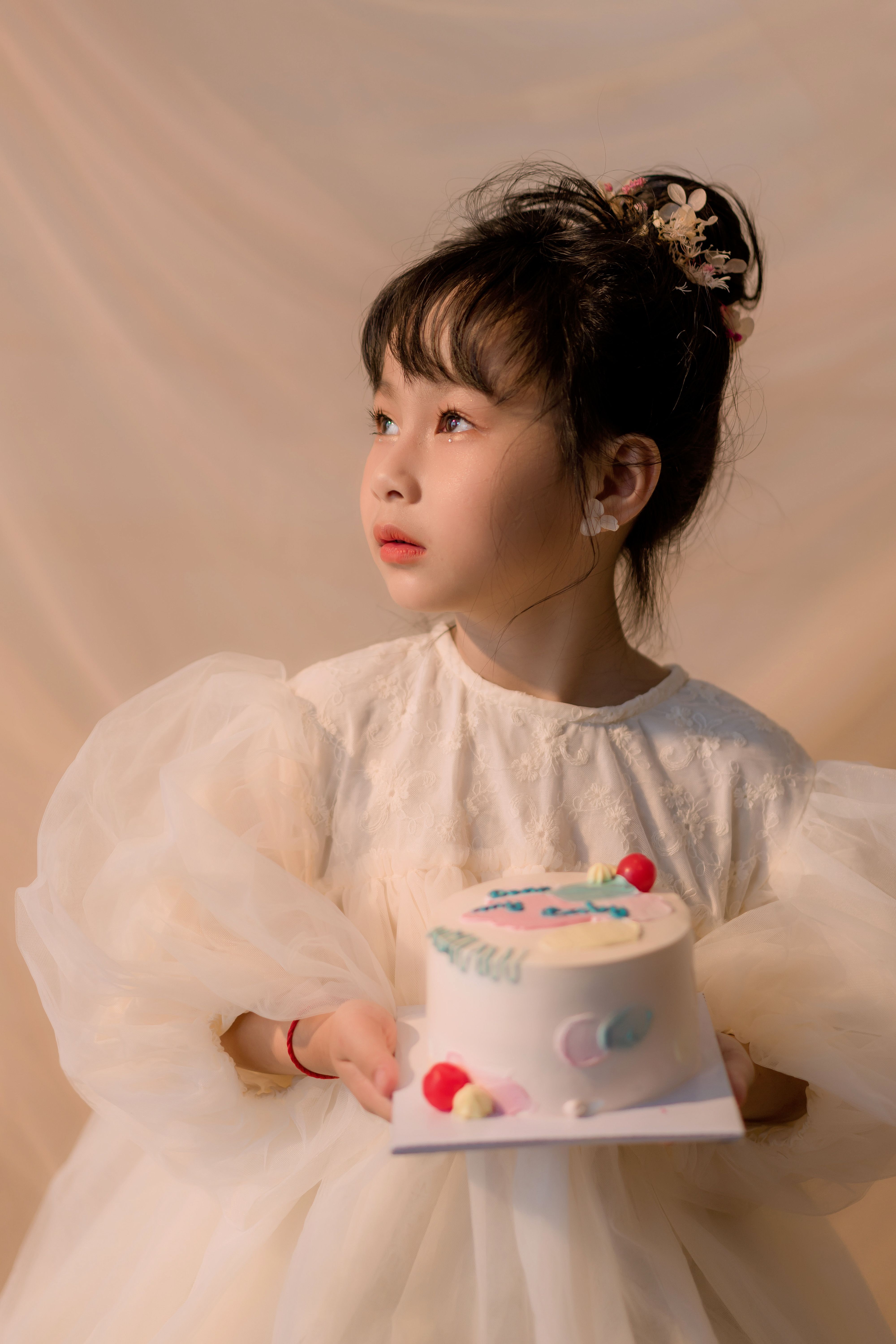 Adorable child in a white dress holding a cake, looking thoughtful indoors.